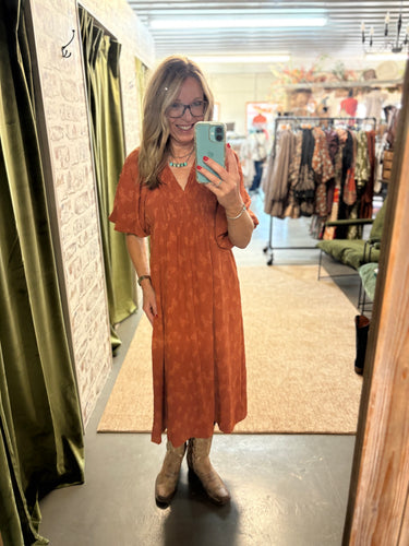 Woman wearing a rust-colored printed midi dress with boots, taking a mirror selfie in a boutique.