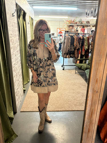 Woman wearing a floral tan dress with boots, posing in a boutique fitting room and taking a mirror selfie.