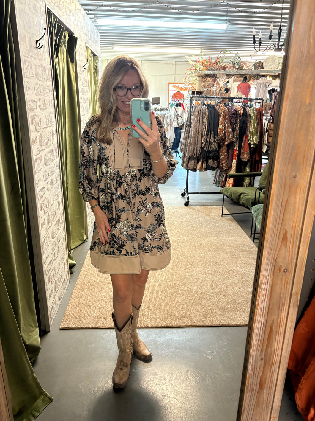 Woman wearing a floral tan dress with boots, posing in a boutique fitting room and taking a mirror selfie.