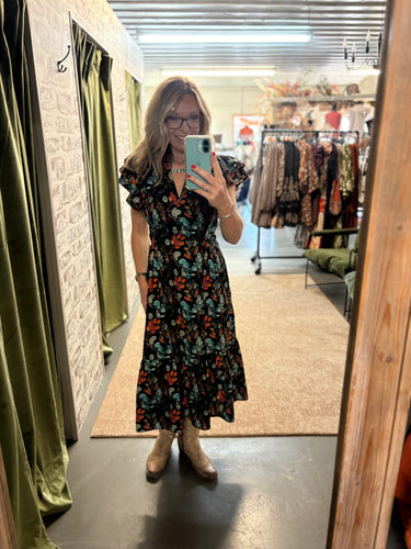 Woman in floral ruffle-sleeve midi dress and boots taking a mirror selfie in a boutique fitting room.