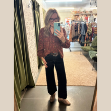 Load image into Gallery viewer, Woman wearing a rust-colored lightweight blouse with a floral print and V-neck, paired with dark wide-leg jeans and woven slip-on shoes, standing in a boutique.
