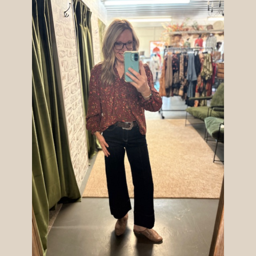 Woman wearing a rust-colored lightweight blouse with a floral print and V-neck, paired with dark wide-leg jeans and woven slip-on shoes, standing in a boutique.