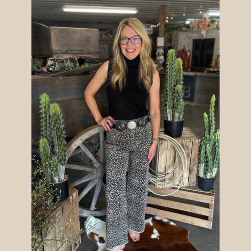 Woman wearing a sleeveless black knit mock neck top styled with leopard print jeans and a concho belt in a western boutique setting.