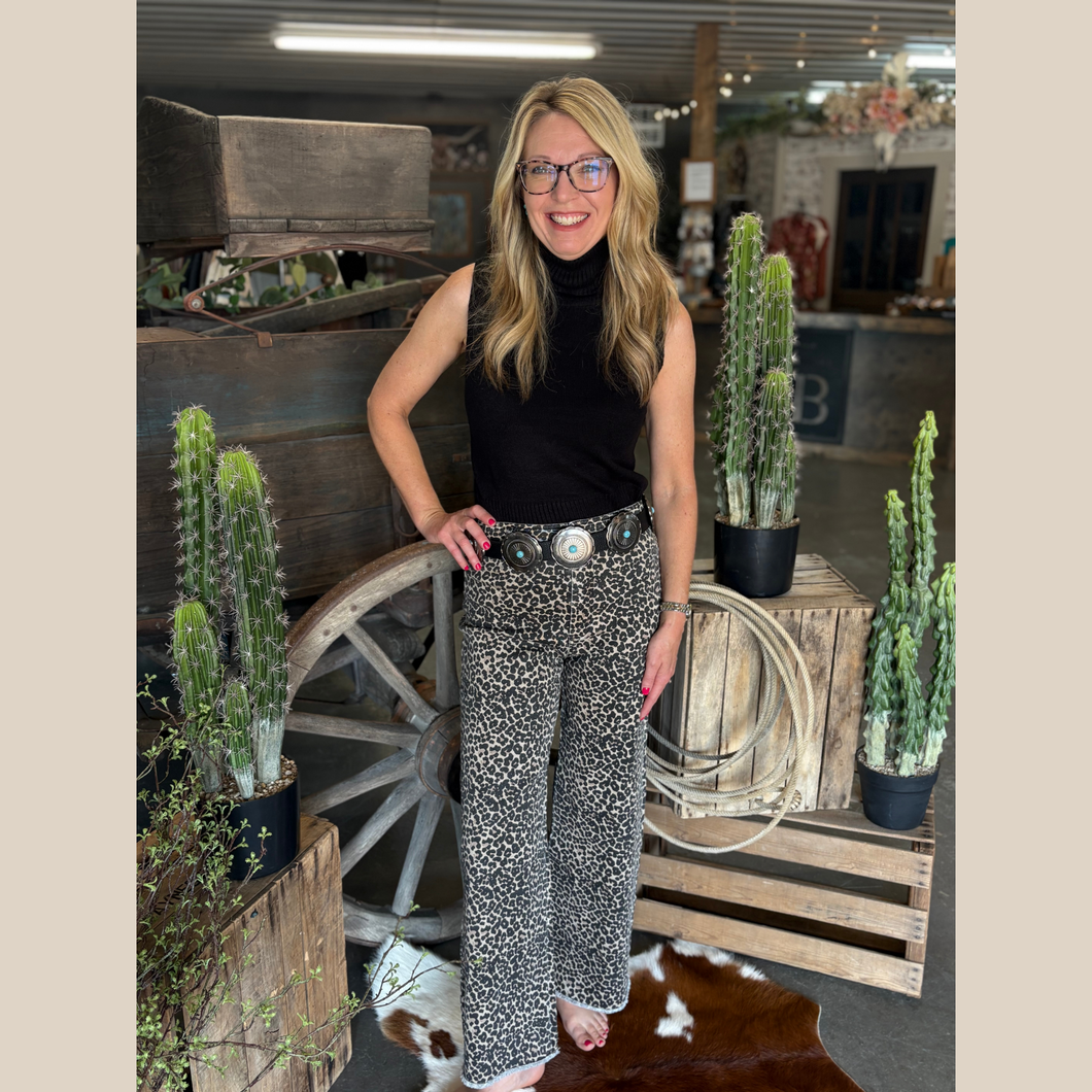 Woman wearing a sleeveless black knit mock neck top styled with leopard print jeans and a concho belt in a western boutique setting.