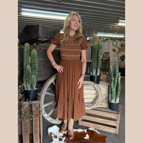 Woman wearing a brown smocked midi dress with contrast stitching, styled with boots in front of a rustic wooden wagon.