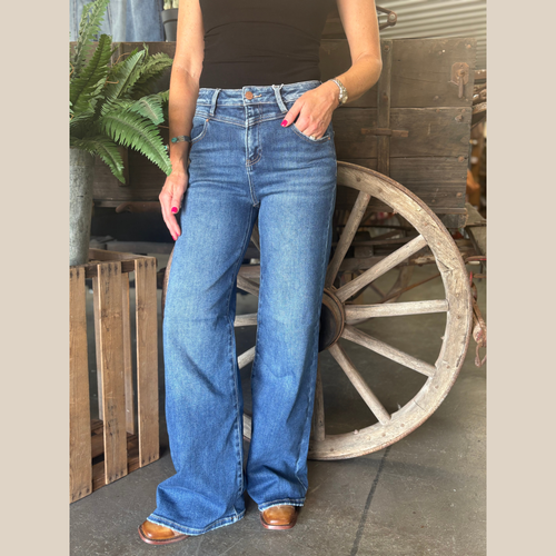 Woman wearing high rise wide bootcut jeans in a dark wash with yoke detail, standing by a vintage wagon wheel.