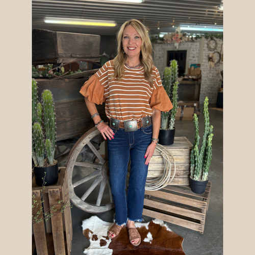 Woman wearing a cinnamon striped puff sleeve top with horizontal stripes, standing indoors near rustic decor.