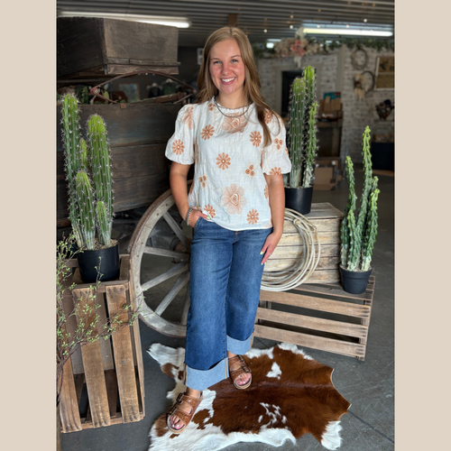 Woman wearing a cream blouse with rust floral embroidery and puff sleeves, standing by cacti and a wooden wagon wheel on a cowhide rug.