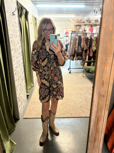 Woman wearing a floral mini dress with ¾ sleeves and boots, taking a mirror selfie in a boutique.