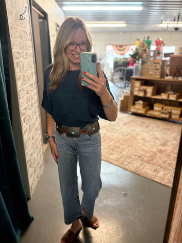 Woman wearing a teal dolman sleeve sweater tucked into light denim with a concho belt and brown sandals.