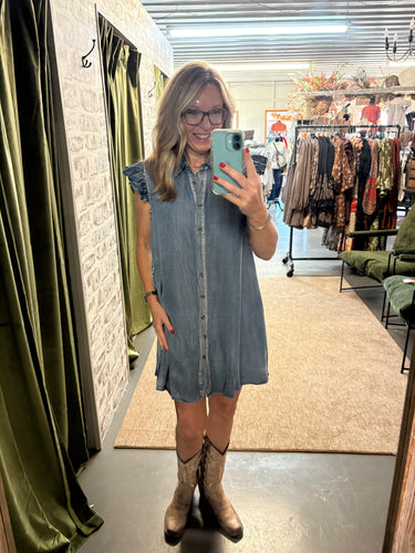 Woman wearing sleeveless button-down denim dress with ruffle sleeves and boots, posing in boutique.