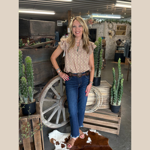 Woman wearing a floral print ruffle sleeve top, standing indoors in front of cacti and a rustic wooden wagon wheel.