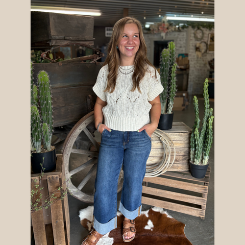 Woman wearing an oatmeal cable knit sweater and wide cuff jeans, standing by rustic decor with cacti and a wooden wagon wheel.