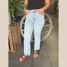 Load image into Gallery viewer, Woman wearing high rise, light wash jeans with a straight leg fit and frayed front hem, standing in front of a rustic wagon wheel.

