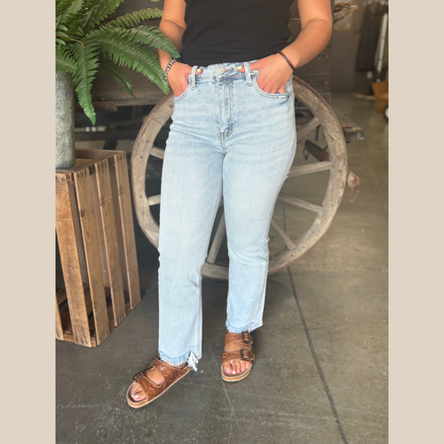 Woman wearing high rise, light wash jeans with a straight leg fit and frayed front hem, standing in front of a rustic wagon wheel.