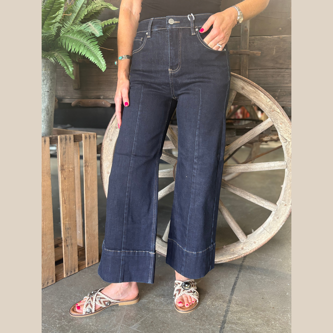 Woman wearing dark wash jeans with front seam and wide cuff, standing in front of a rustic wooden wagon wheel.