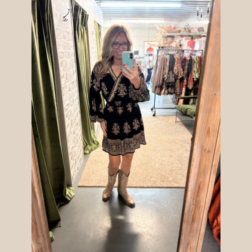 Woman wearing The Sable Trail Dress, a black long-sleeve dress with tan floral print, V-neckline, and elastic waist, styled with cowboy boots.