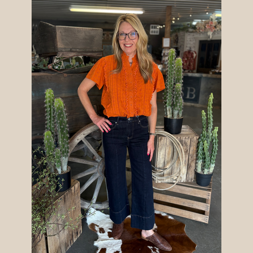 Bright orange eyelet short-sleeve top with ruffle trim and mock neck, paired with dark wide-leg jeans in a western boutique.
