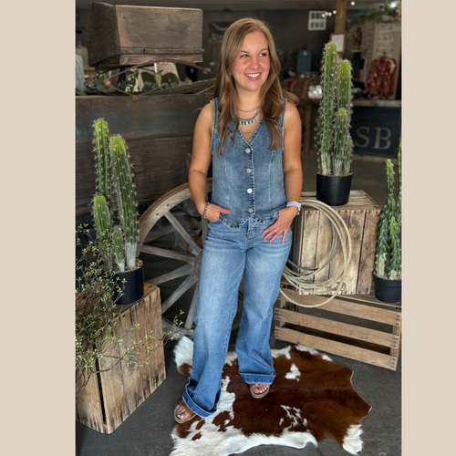 Woman wearing a sleeveless denim v-neck button down vest and wide-leg jeans, standing in a rustic western boutique with cactus and cowhide decor.