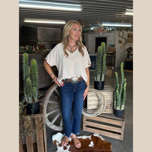 Woman wearing a V-neck blouse with contrasting whipstitch detail, shown in oatmeal and mocha colors, styled in a western indoor setting.