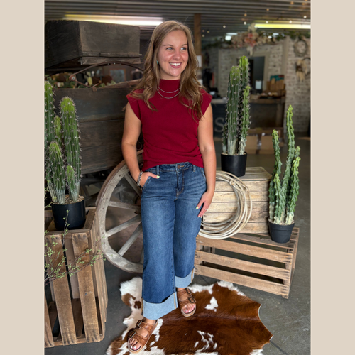 Woman wearing Wine Ruched Mock Neck Top with wide cuff jeans, standing by cacti and a wooden wagon wheel on a cowhide rug.